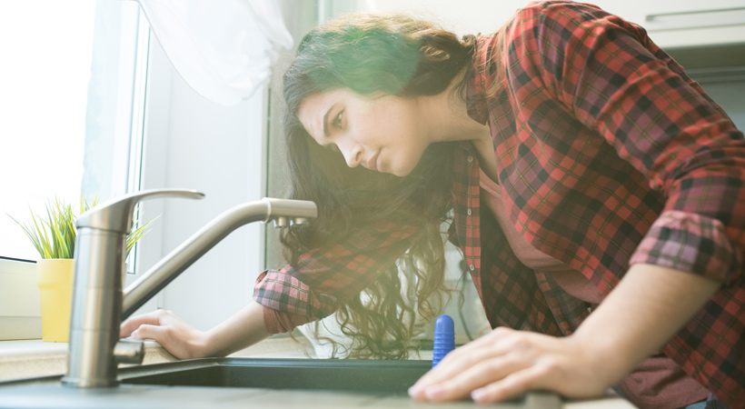 A woman in a red plaid shirt inspects a kitchen sink, leaning in with a focused expression. Sunlight streams through a nearby window, illuminating a potted plant.