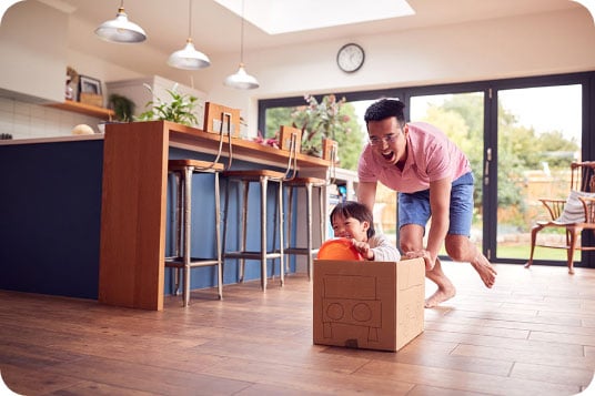 Father playfully pushes his young daughter in a cardboard box across a bright, modern kitchen floor.