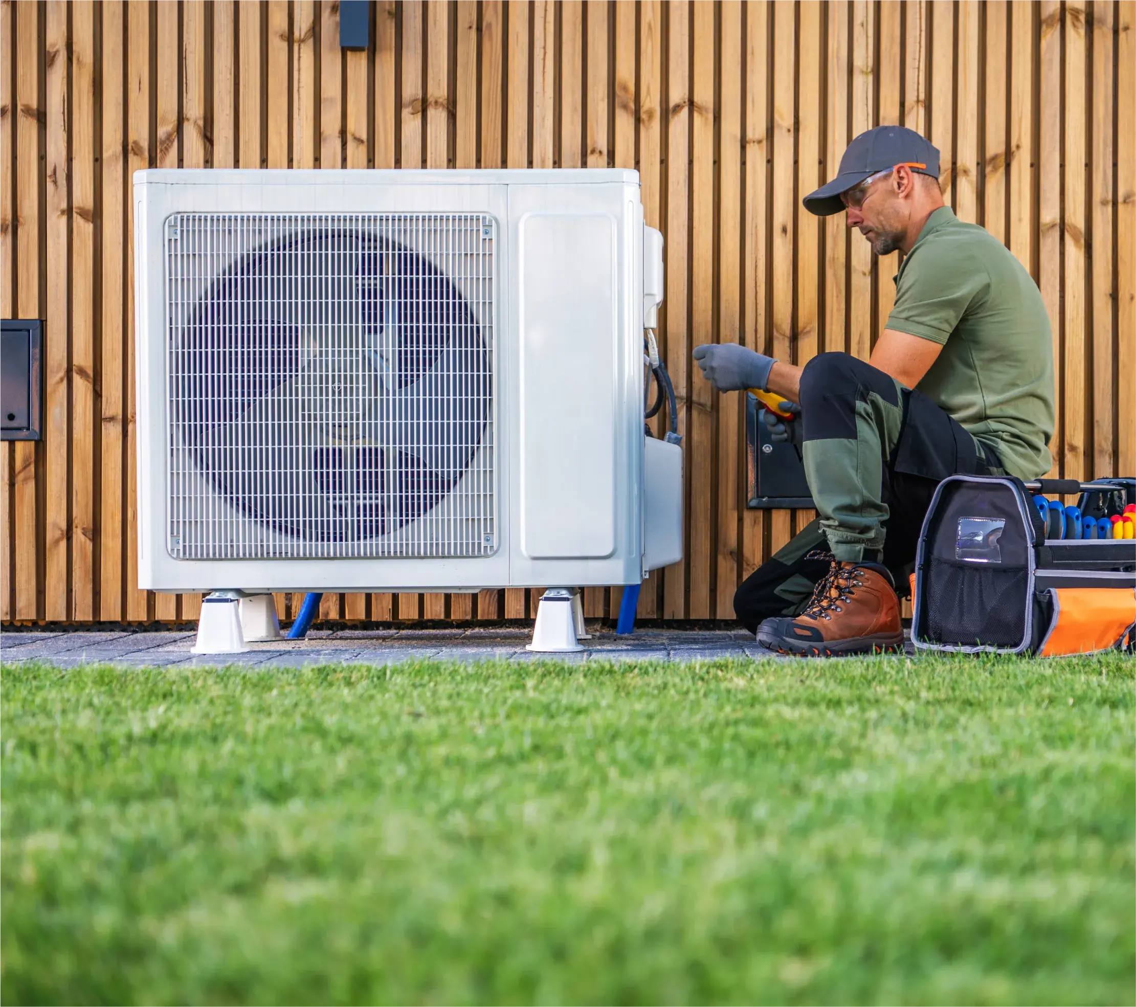 Technician working on an outdoor HVAC unit beside a wooden wall, with tools and equipment laid out on the grass