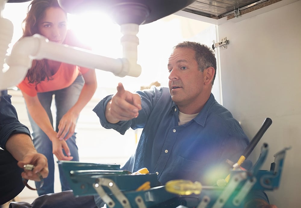 Plumber pointing to pipes beneath a kitchen sink while a homeowner looks on
