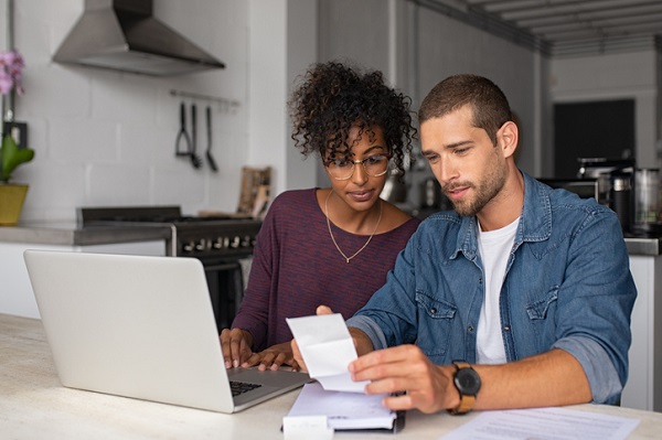 Young multiethnic couple checking bills while managing accounts on home banking app. Serious guy and african woman sitting at home discussing finance for the month. Young casual man and girl using laptop while looking at invoice and plan the budget to save, wondering if a home warranty is worth it.