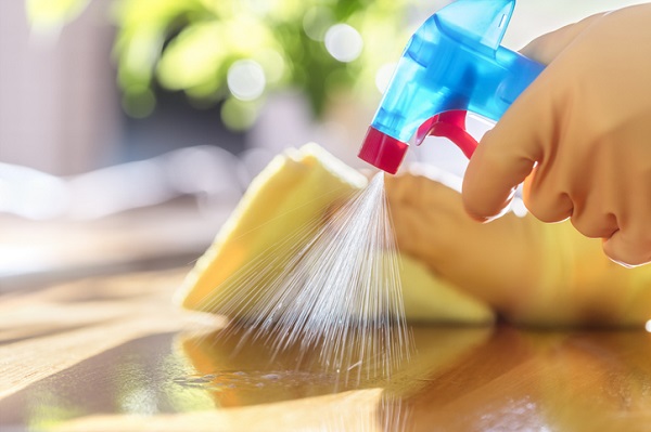 A blue spray bottle with a red trigger and tip spraying liquid on a wood surface. A White person's hand is holding the spray bottle in the left hand and a yellow microfiber cloth in the right.
