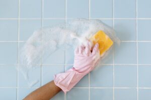 A light blue tile being washed down with soap and a yellow sponge. A brown-skinned arm with a pink glove is doing the cleaning.