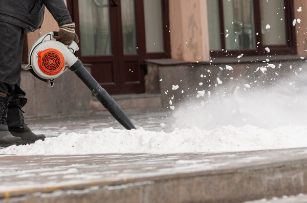 A person's arm using a gray and orange leaf blower to clear snow from a sidewalk