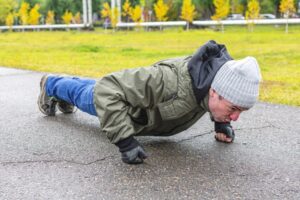 Man in winter clothes doing pushups with balled fists on a sidewalk near a park. 