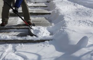 A person shown from the legs down using a black handled, gray shovel to remove snow from a set of stairs that are long on length but short in height during a sunny day