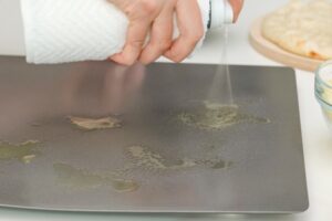 A person's hand gripping a cooking-oil spray can and spraying oil on a square, metal surface with pizza dough in the background
