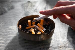 A white caucasian hand holding a cigarette whose ashes are tapped into an ashtray.