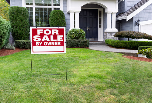 A nice white house with a FOR SALE BY OWNER sign in front of it.