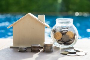 A wooden-model house next to a coin jar with coins in it. There are also coins in front of the house. A pool is in the background.