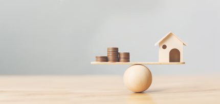 A tiny model house and a stack of quarters balanced on a see-saw atop a wooden ball