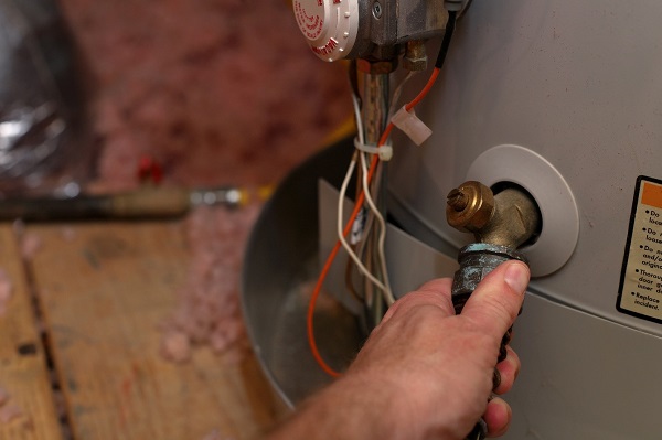 A White person's left hand gripping the spigot of a water heater. The tank is gray, and the spigot is brass with signs corrosion.