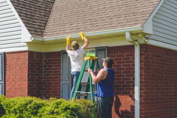 A White woman in blue jeans, a white t-shirt, yellow gloves, and a tan hat on a green A-frame ladder cleaning out gutters. A White man in a blue tank top and gray hat is holding the ladder. The house is red brick with white siding.