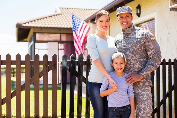 A White woman in a three-quarter length light blue shirt and blue jeans, a black man in Army fatigues including a hat, and a younger girl in a light-purple shirt and blue jeans all stand in front of a wood-picket fence. In the background is a ranch-style house with and American flag