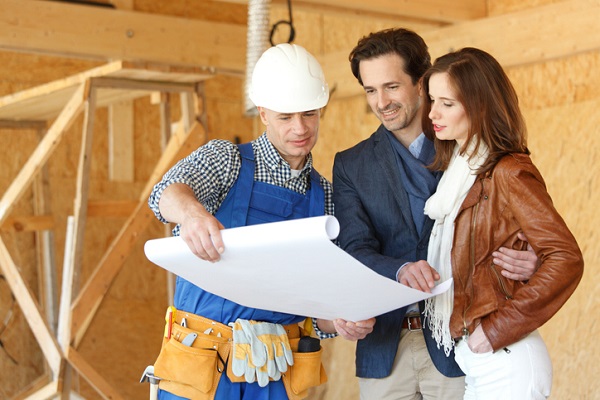 A White builder in a white hardhat shows blueprints to a White man in a suit coat and khakis and a White woman in a brown leather jack and white pants.