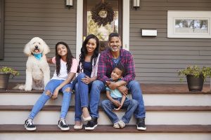 A husband, wife, two daughters, and big white fluffy dog sitting on a front porch smiling.