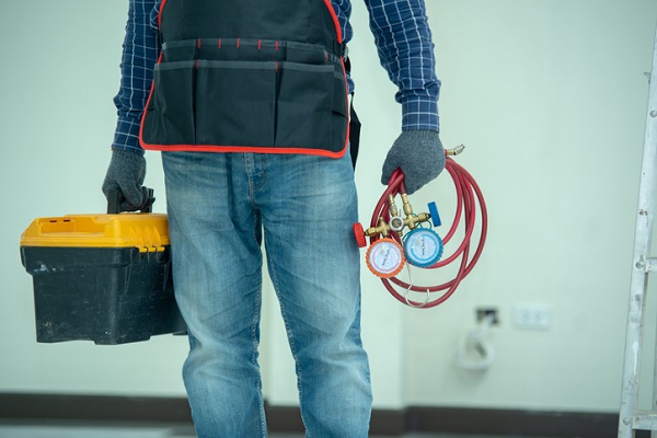 A picture of a man in jeans and a flannel with a work apron over the flannel. he's wearing gray gloves and holding a toolbox with a yellow top in his right hand, In his left hand is an air conditioner coolant gauge. You can only see his legs and part of his torso.