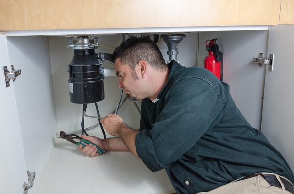A Hispanic man in a dark blue shirt and pliers in his right hand lying next to a black garbage disposal under a sink.