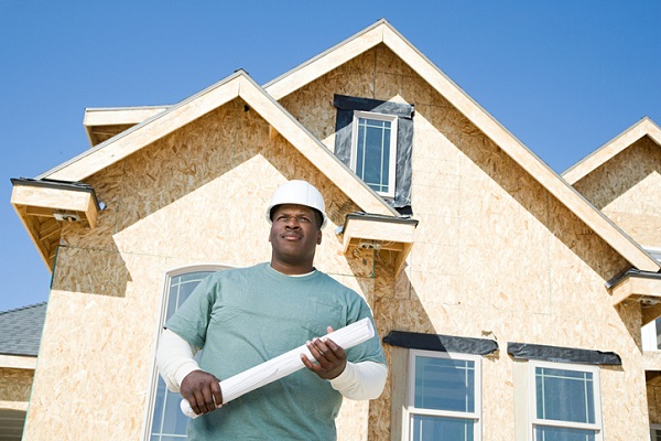 A Black home builder in a white hard hat standing in front of a wood-frame house with rolled up blue prints in his hand.