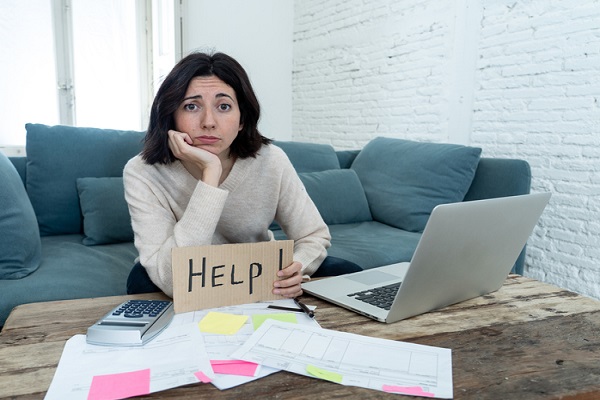 A young White woman with shoulder-length black hair wearing a long-sleeved tan sweater sitting on a blue-gray couch. She's holding a small cardboard sign that says Help with her eyebrows slightly furrowed and her head resting in the palm of her right hand. On the table in front of her are scattered papers, a calculator, and an open laptop