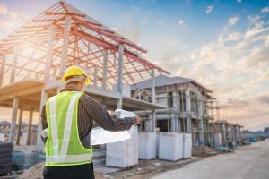 A construction worker in a high-visibility vest standing in front od a wood-frame house that's being built.