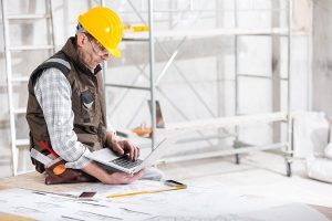 A White construction worker sitting at a white table in front of scaffolding. He's looking at a laptop.