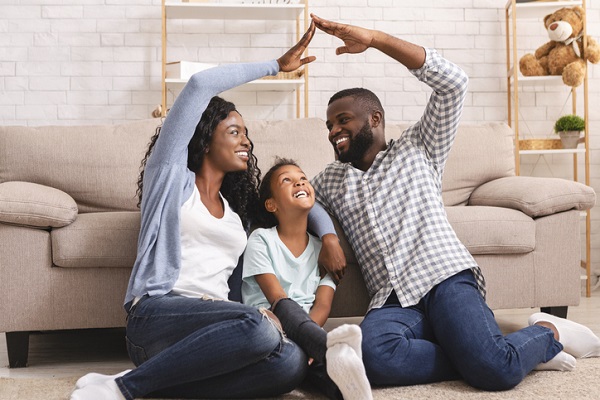 A Black man and woman sitting next to each other on the floor smiling and looking at each other. They are making the shape of a roof of a house over their young daughter. Everyone is happy.