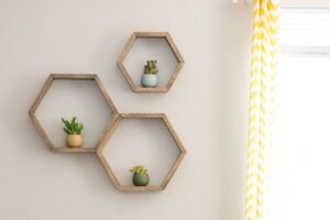 Three wooden, hexagonal shelves on a beige wall, each holding one potted succulent plant, next to a window with a tangerine and white striped curtain