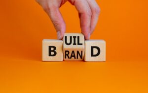 Three wooden blocks. The one on the left has a B on it. The one on the right has a D on it. The one in the middle has UIL on one face and RAN on another. A hand is holding the middle block so that it spells out Build Brand, depending on how you look at it.