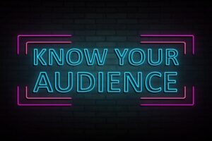 The phrase KNOW YOUR AUDIENCE on a black background. The letters are turquoise and surrounded by a magenta border. It looks like a neon sign,