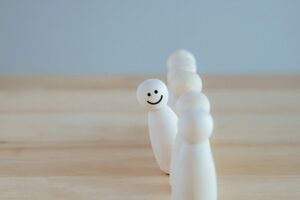 A row of white, wooden pegs that represent people. One of the wooden pegs has a smiley face drawn on it and is standing out from the crowd.