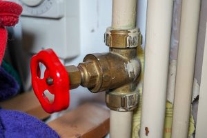 Three light tan pipes against a wall. the left-most pipe has a brass valve with a bright red wheel on it