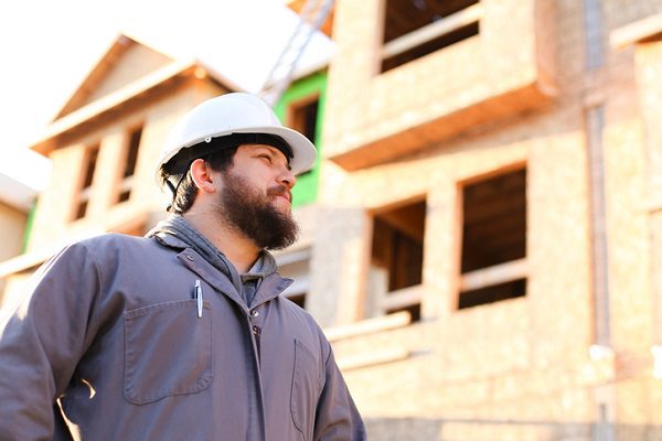 A White man with brown beard and white hardhat and gray zip-up jacket standing in front of the construction framing of a house