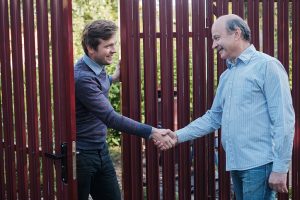 A White man with full brown hair wearing a light blue collared shirt under a blue sweater, along with clue jeans, shaking hands with an older, balding man with gray hair and a mustache, who is wearing a light blue, long-sleeved collared shirt and blue jeans. They are standing in front of a burgundy-colored fence with a door that's partially open