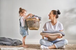 A White woman sitting on the ground cross-legged holding fresh and folded towels. On her left, a White child in overalls is handing her a wicker basket of laundry, Both are smiling. It is a happy moment in history.