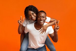 A Black man with short, cropped hair giving a piggyback ride to a Black woman with braces and long wavy hair. They're both in blue jeans and white t-shirts, smiling, and giving a peace sign, the man with his left hand and the woman with her right hand.