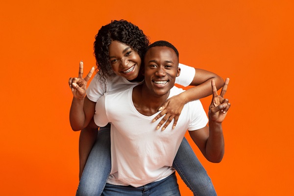 A Black man with short, cropped hair giving a piggyback ride to a Black woman with braces and long wavy hair. They're both in blue jeans and white t-shirts, smiling, and giving a peace sign, the man with his left hand and the woman with her right hand.