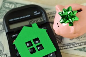 An old-school calculator with the word BONUS on its display. A green paper cutout of a house rests on the keypad. A piggy bank with a green bow is to the right of it. Everything is on a pile of money.