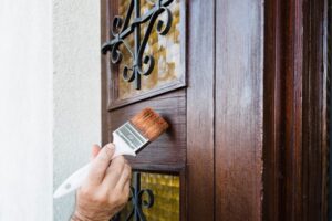 Worker hand paints wooden front door with paintbrush. The door is a dark wood with a pane glass window.