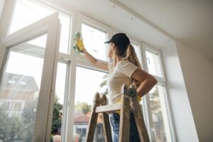 A white woman in blue jeans, white t-shirt, yellow gloves, and black hat standing on a small wooden ladder, cleaning interior windowpanes with a green rag on the inside of a white-walled house