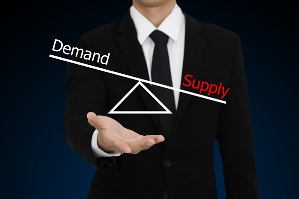 The torso and right hand of a White business man in a black suit, white shirt, and black tie. His hand is balancing a fulcrum. The see-saw on the fulcrum has "demand" on one side and "supply" on the otherr. The "Supply" side is pointing downward, signifying more supply than demand