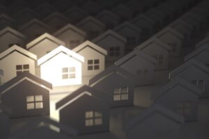 A group of about 50 small, white cardboard cutouts of houses. Each house cutout has a four-pannel window on the right side. One house is bright white and under a spotlight, while the other houses fade into a dark background. The farther away from the spotlit house they are, the more in the shadows they are,.