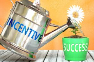A large metal watering can on the left with the word "incentive" printed on it in blue. The can is watering a white daisy that 's planted in a ime green pot that has the word "success" on it in white letters. The can and pot are on a wood plank patio in front of an orange background