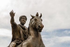 The statue of Marcus Aurelius, Campidoglio, Rome, Italy. Marcus is riding his horse with his right arm stretched outward and his fingers relaxed. He has curly hair and a curly beard. The statue is bronze
