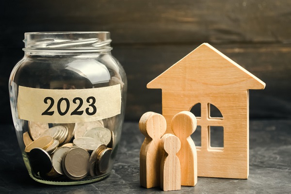 Three small wooden figurines of people, just the head and torso, representing parents and a child in front of a flat, wooden model of a house with an arched window with a cross in the middle of it. To their left is a jar with coins in it. On the jar is a piece of tan masking tape with 2023 printed on it,.