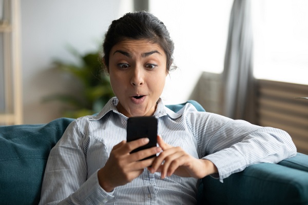 Young Indian woman in a blue and white striped, long sleeve shirt sitting on a couch. She's holding a smart phone and looking at it in with a "pleasantly surprised" look--eyebrows raised, an open-mouth smile