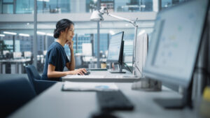 An Asian woman in a dark blue shirt sitting at a desk in front of a computer holding a computer mouse. She's in a sleek office that has lots of glass and steel