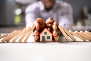 A small wooden model of a house surrounded by a pair of Black hands. The hands are protecting the house from two rows of dominos that are falling around it on each side.