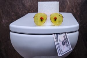 A toilet with two $50 bills sticking out from the between the rim and lid so that it looks like a tongue. On top of the lid are a pair of sunglasses with fake eyes, and behind the glasses is a roll of toilet paper