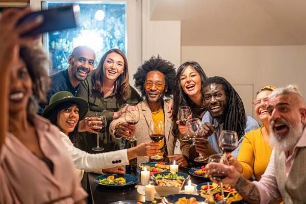 Multigenerational, multiracial group of people at a dinner table toasting with glasses of red wine. A Black woman in the foreground is taking a selfie of everything.